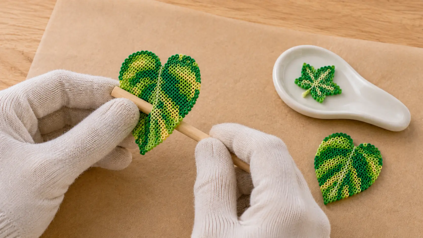 Gloved adult hands gently shaping a connected warm pothos fuse bead leaf over a wooden dowel beside a curved ivy leaf